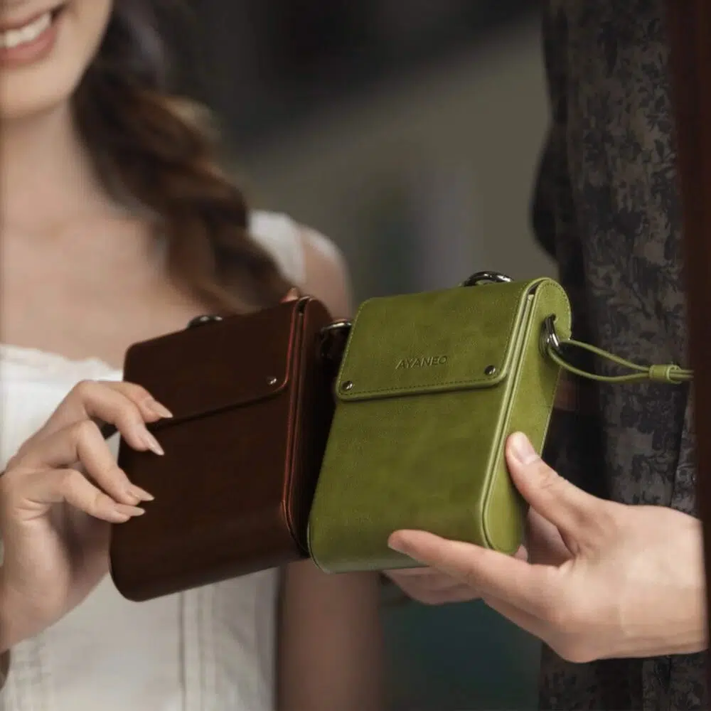 Dois indivíduos seguram sacos de pele compactos. Uma bolsa é verde e a outra é castanha, ambas com o logótipo AYANEO e um design elegante. O cenário sugere uma utilização casual ou profissional.