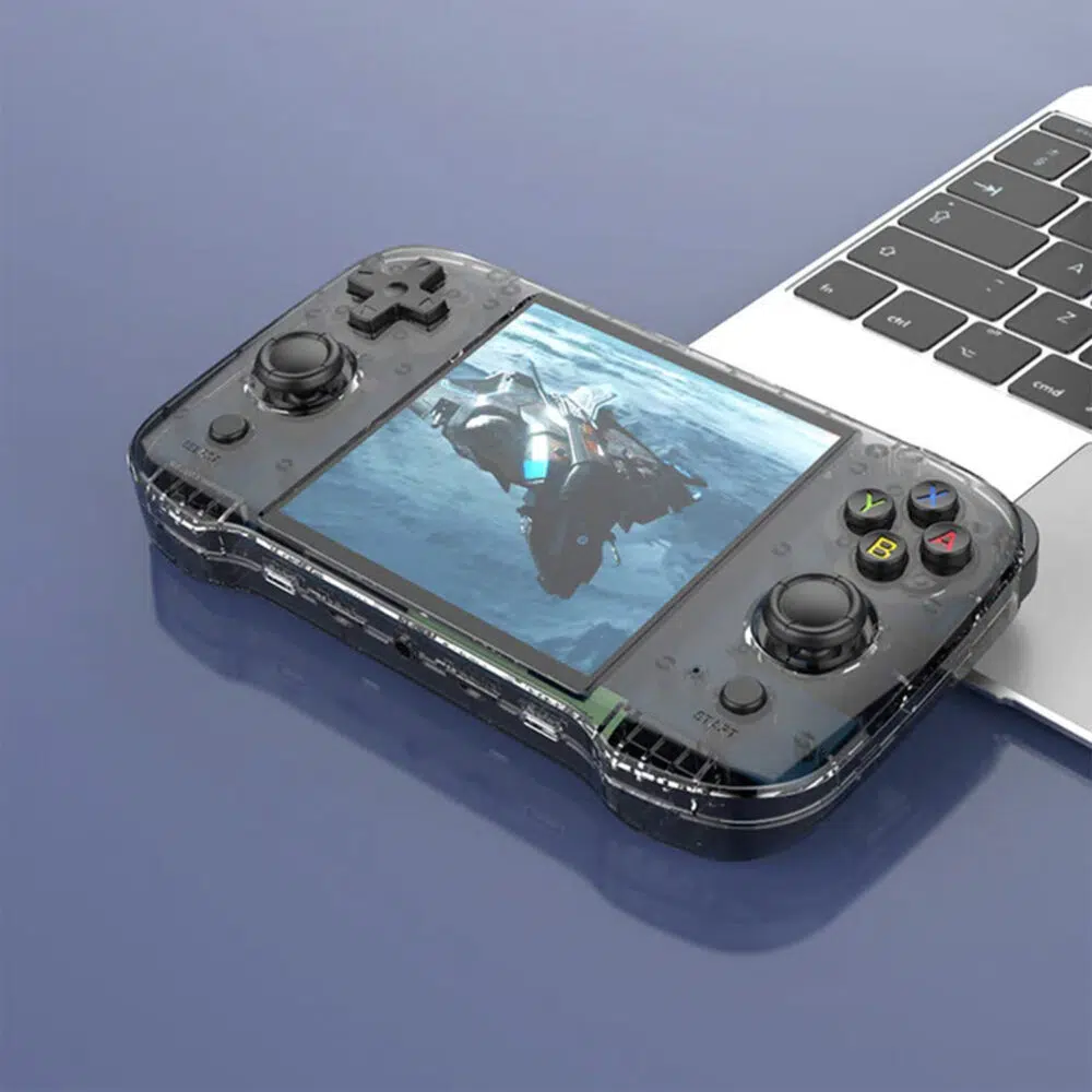 Un dispozitiv portabil negru transparent pentru jocuri, plasat pe un birou lângă un laptop, care afișează pe ecran o navă spațială futuristă.