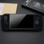 A black AYANEO 3 handheld gaming console lies centered on a dark surface, featuring a newly applied tempered glass screen protector. The console's dual joysticks are illuminated with circular white LED rings. To the top right, the product packaging is visible, showing a black box labeled "METIK SCREEN PROTECTOR" with an illustrative diagram of the installation process. The sleek, matte black finish of the device and the glossy reflection on the protected screen create a high-end, tech-focused aesthetic.