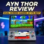 The image features a dual-screen, clamshell-style handheld gaming device centered on a dark surface against a blurred, neon-lit gaming room background. The device is grey with colorful "ABXY" buttons and glowing blue rings around the joysticks. The top screen displays a vibrant 3D Sonic the Hedgehog game, while the bottom screen shows a stylized menu or secondary game interface.