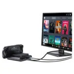 A black GPD WIN 5 handheld console is shown docked and connected via cables to a monitor. The monitor displays the Steam OS-style interface with a grid of game titles such as Dead Cells, Deathloop, Portal 2, and Stardew Valley, illustrating the device's ability to drive a desktop gaming experience.