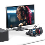 A desk setup showing the GPD WIN 5 docked and connected to an external monitor via cables. The same image of a female character with pink hair is displayed on both the small console screen and the large external monitor, demonstrating the dock's video output capability.