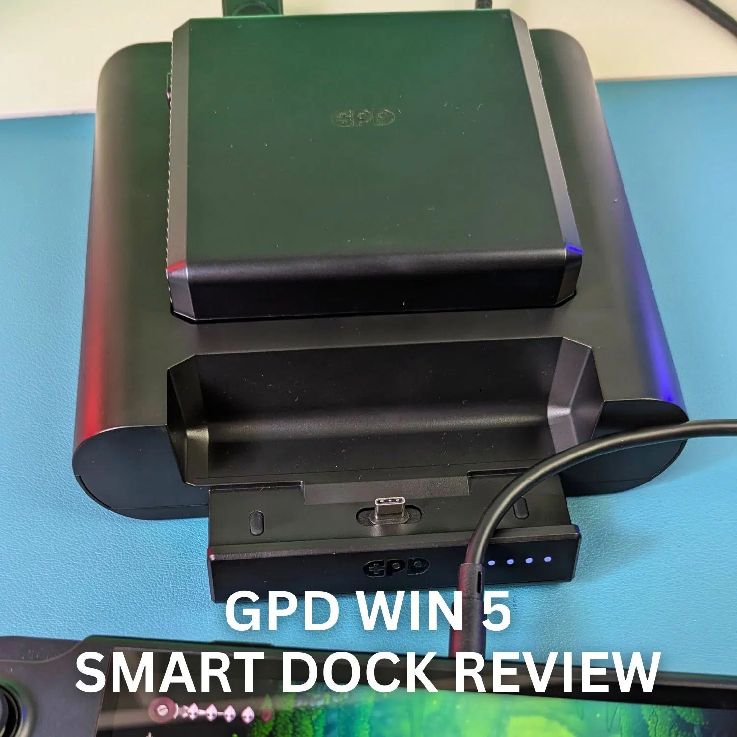 A close-up photograph of the black GPD WIN 5 Smart Dock sitting on a light blue surface. The dock is designed to hold the GPD WIN 5, with the console's screen partially visible in the foreground, displaying a gaming interface. The dock itself is connected by a black cable, and its LED indicator lights are visible near the charging port. Large white text overlaid on the image reads, "GPD WIN 5 SMART DOCK REVIEW."