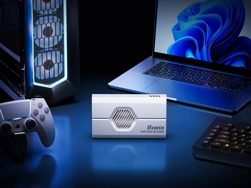 A lifestyle shot of the white SSD reader placed centrally on a blue-lit desk setup, flanked by a gaming PC tower, a game controller, and a laptop.