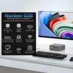 A lifestyle render of the Nucbox G3S mini PC situated on a white desk with a keyboard, mouse, and a monitor displaying an abs